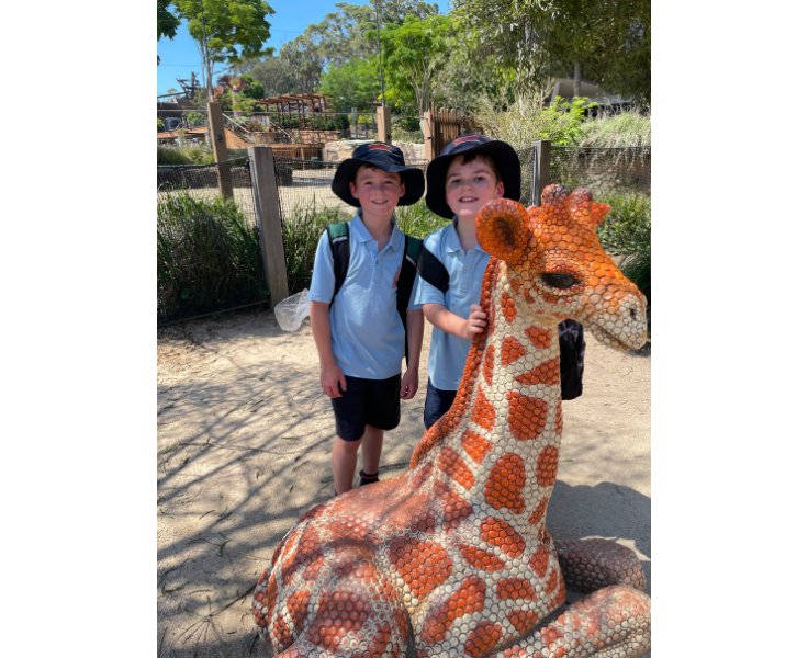 Two children at taronga zoo smiling next to a statue of a giraffe